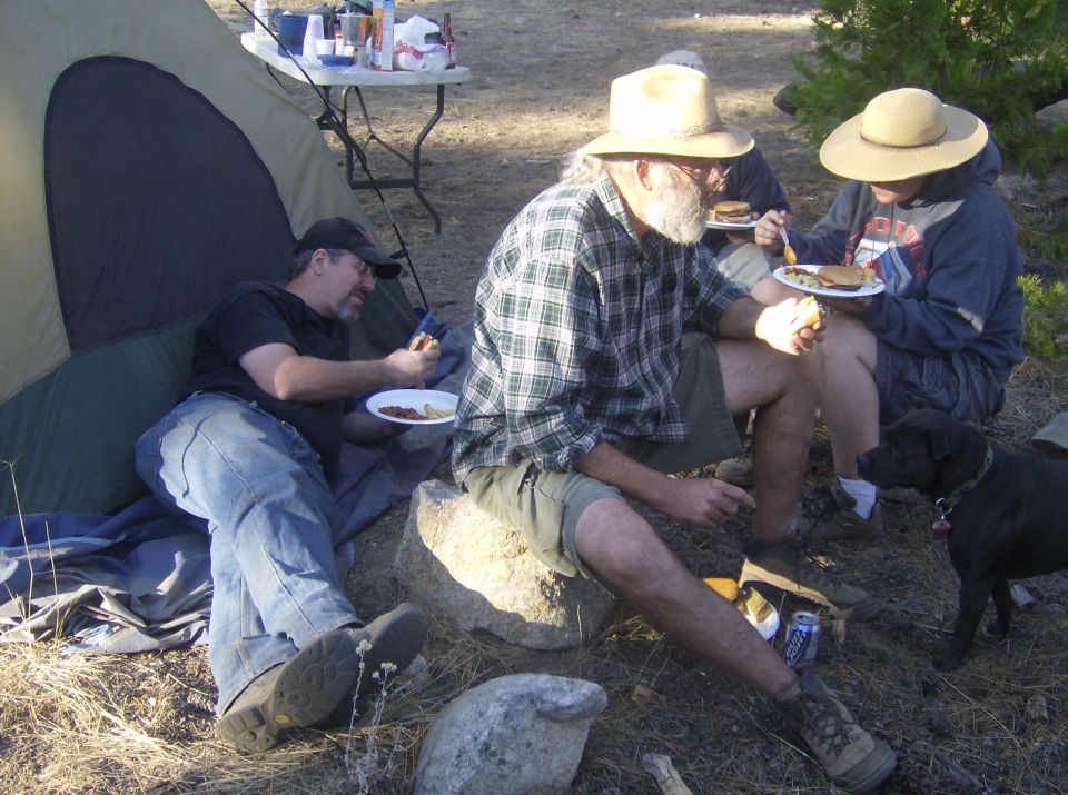 People eating at a campsite next to a tent, with a dog nearby