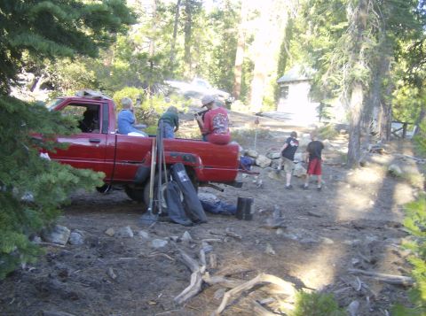 1986 Ford Bronco - People are gathered around a red pickup truck in a wooded area, with some sitting in the truck bed.