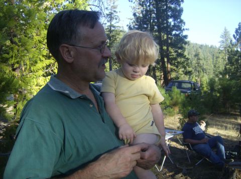 1986 Ford Bronco - An older man in a green polo holds a blond toddler in a yellow onesie at a sunlit pine-forest campsite, with another person seated in a cam…