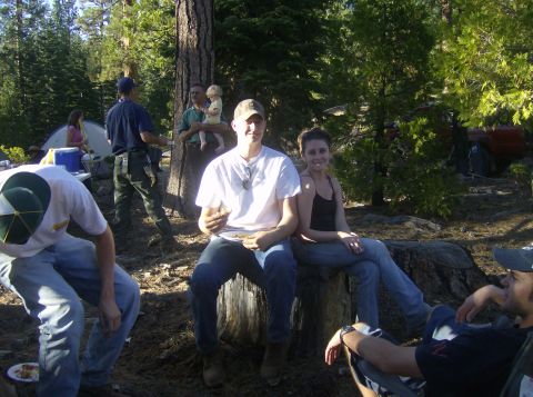 1986 Ford Bronco - Group of people relaxing and eating at a pine-forest campsite, with a couple seated on a large tree stump in the foreground and a dome tent…