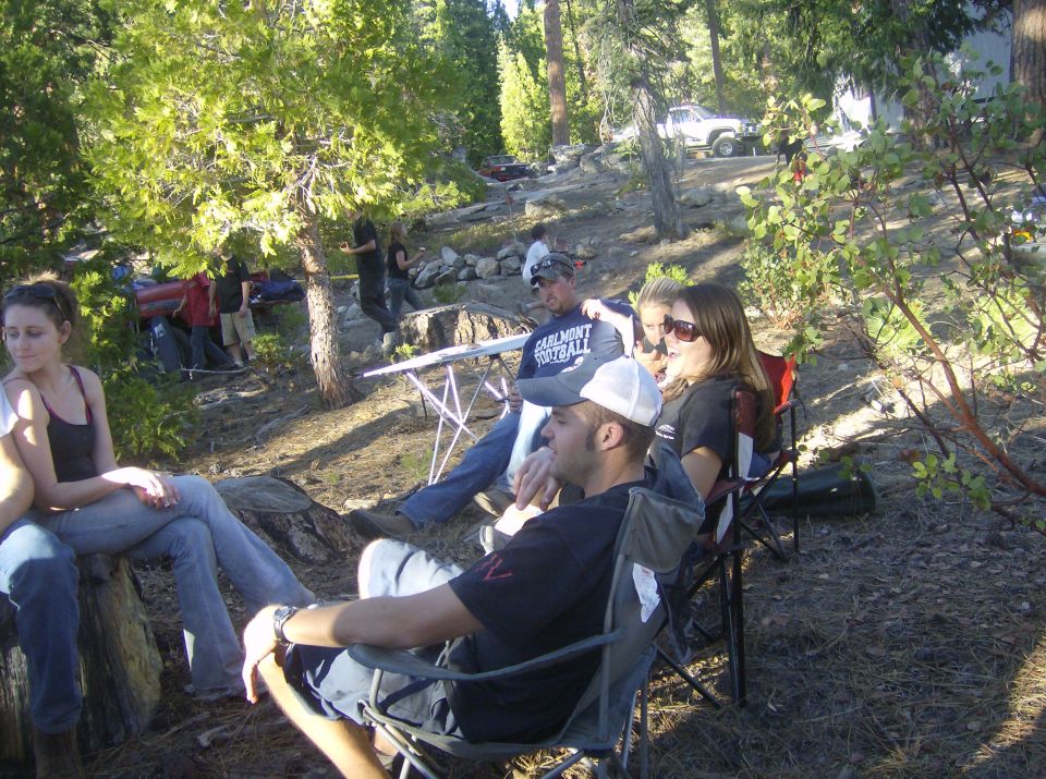 Group in camp chairs at a pine forest camp with trucks parked on the slope behind