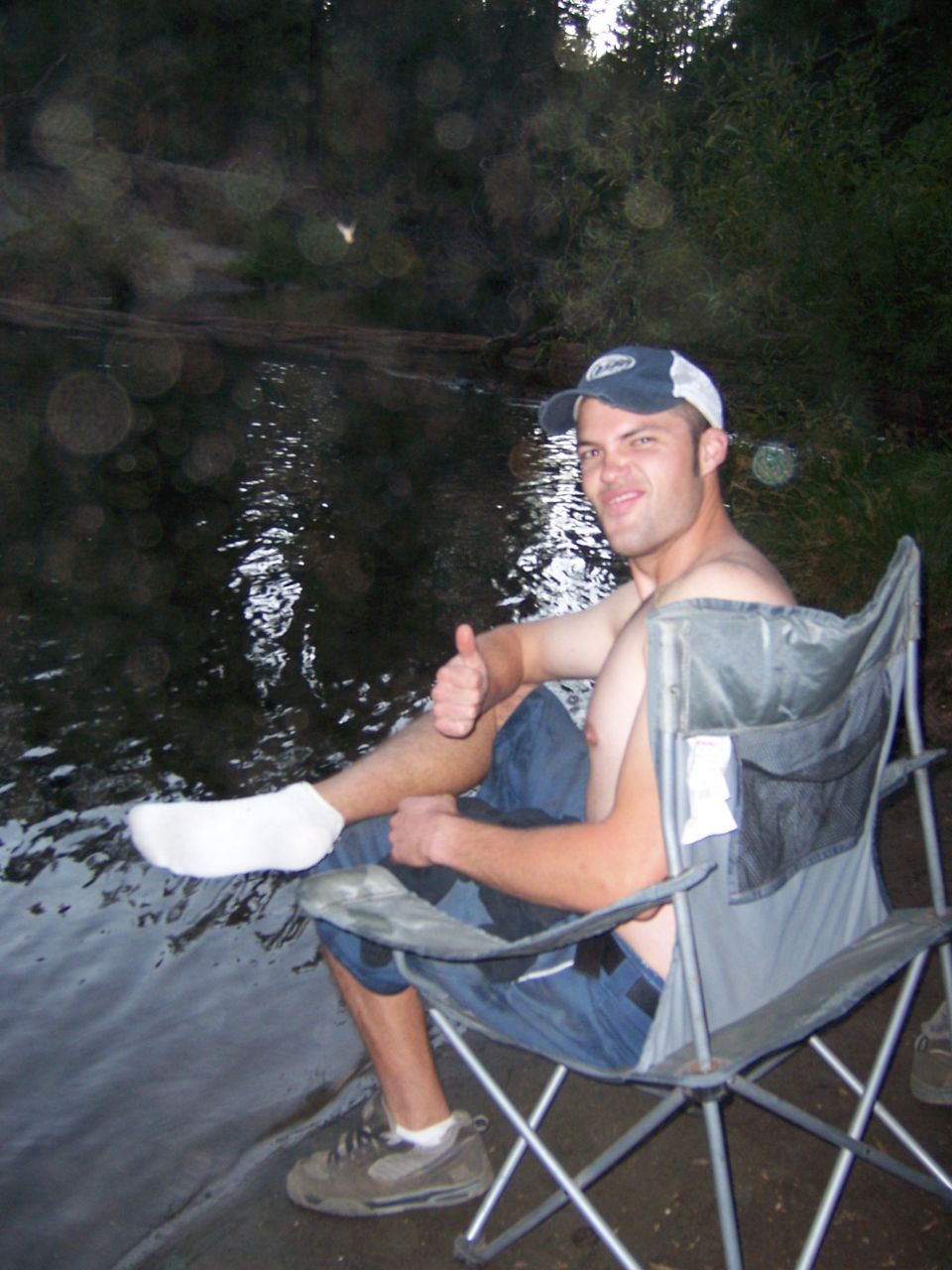 Man in a camping chair gives a thumbs up next to a river, wearing a baseball cap and jeans