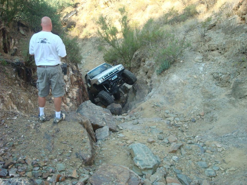 1986 Toyota Hilux modified for off-roading, climbing over rocks in a steep, dry ravine with a man watching