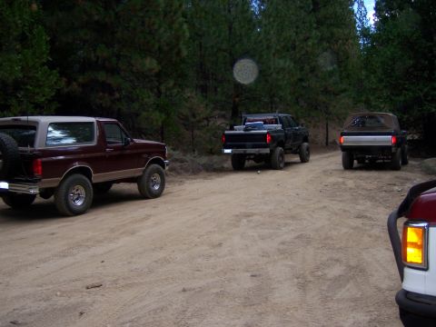 1991 Ford Bronco - Three trucks are parked on a dirt road surrounded by trees.