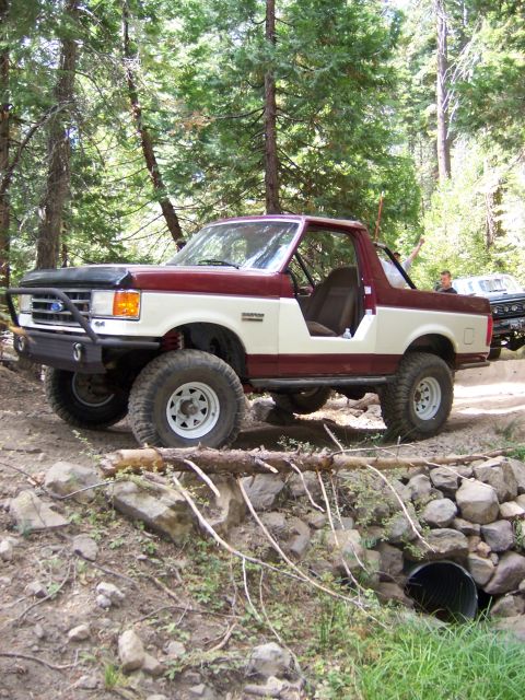 1991 Ford Bronco - A modified Ford Bronco navigates a rocky trail in a wooded area, with another vehicle visible behind it.