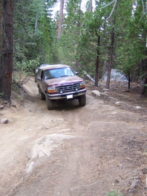 1991 Ford Bronco - A maroon Ford Bronco navigates a rocky dirt path surrounded by trees.