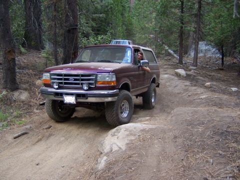 1991 Ford Bronco - A maroon Ford Bronco navigates a rocky dirt trail through a forest.