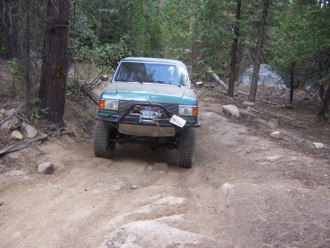 1991 Ford Bronco - This modified Toyota Land Cruiser FJ60 navigates a rocky off-road trail surrounded by pine trees.