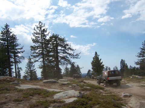 1991 Ford Bronco - Two off-road vehicles traverse a dusty trail through a wooded, rocky landscape under a cloudy sky.