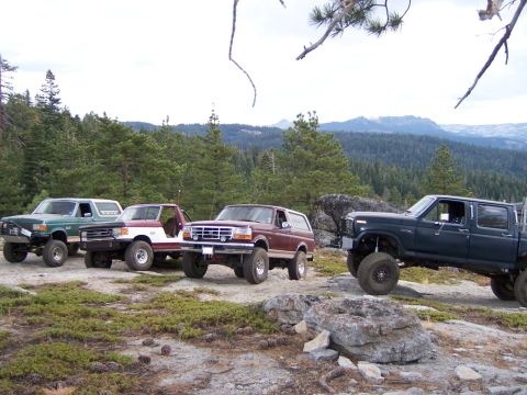 1991 Ford Bronco - A group of modified trucks and SUVs are parked on a rocky incline with a forested mountain range in the background.