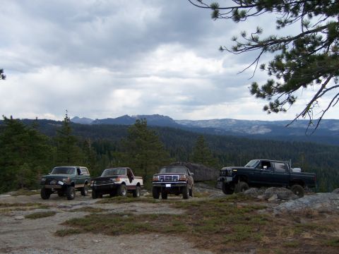 1991 Ford Bronco - Several trucks are parked on a rocky outcrop overlooking a vast forest and mountain range under a cloudy sky.