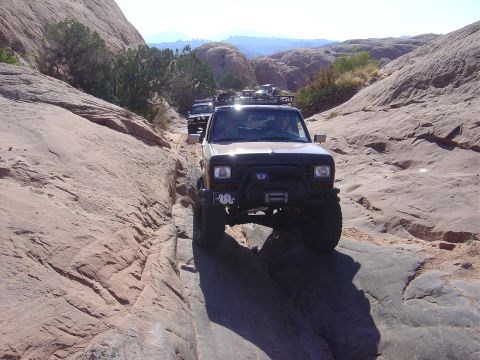 1986 Ford Bronco II - Multiple off-road vehicles, including a 1986 Ford Bronco II, traverse a challenging rocky path in a desert canyon.