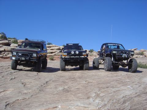1986 Ford Bronco II - Three modified SUVs and trucks are parked on a rocky landscape under a clear blue sky.