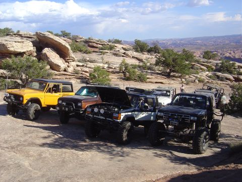 1986 Ford Bronco II - Several modified off-road vehicles are parked on a rocky, desert landscape under a cloudy sky.