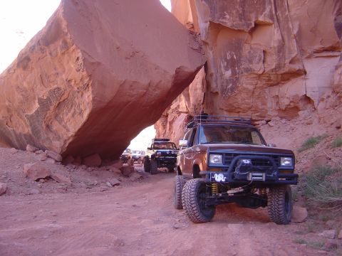 1986 Ford Bronco II - Four-wheel drive vehicles navigate a tight trail under a towering rock formation.