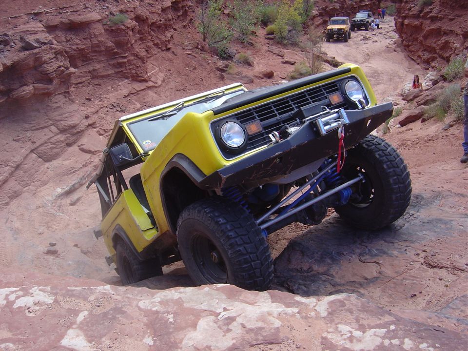 Yellow first-generation Ford Bronco climbing a rocky trail with other vehicles in the background