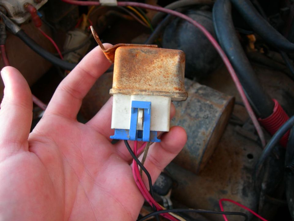 Hand holding a rusty automotive relay with blue plastic connector and wires