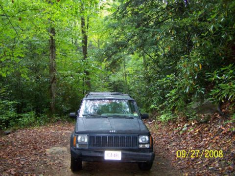 1992 Jeep Cherokee - A dark gray Jeep Cherokee is parked on a dirt road surrounded by dense foliage.