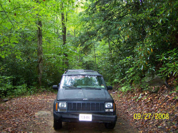 Dark gray Jeep Cherokee parked on a dirt road in a forest, front view