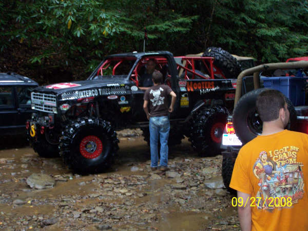 Black modified off-road truck with red roll cage and large tires, parked on a muddy trail