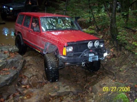 1992 Jeep Cherokee - A red Jeep Cherokee navigates a challenging off-road trail with muddy terrain and large rocks.