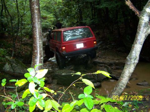 1992 Jeep Cherokee - A red Jeep Cherokee navigates a challenging, muddy off-road trail surrounded by dense forest and rocks.
