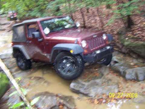 1992 Jeep Cherokee - A red Jeep Wrangler tackles an off-road trail, splashing through water and over rocks, with another vehicle following behind.