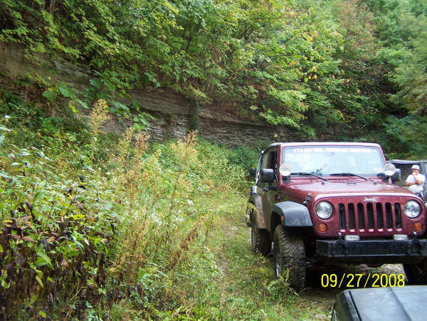 Red Jeep Wrangler parked on a dirt road next to a rocky cliff face. Another Jeep is partially visible behind it