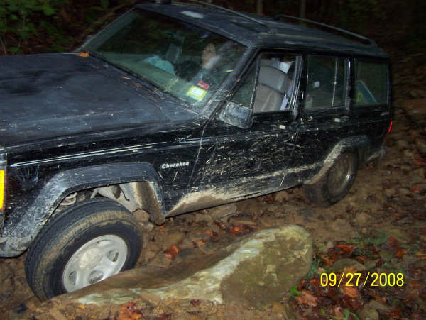Black Jeep Cherokee stuck in mud, front passenger side wheel deep in a rut. Visible "Cherokee" badge