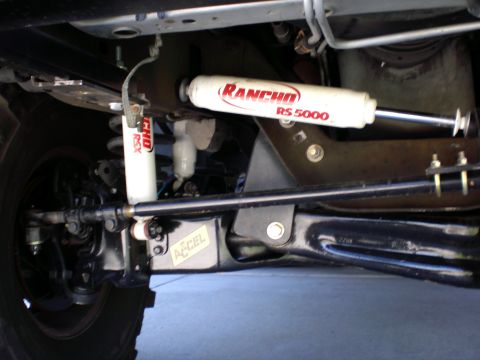 1996 Ford Bronco - Underside view of a truck's front suspension showing two white Rancho RS5000 shock absorbers with red lettering, a black steering stabilize…