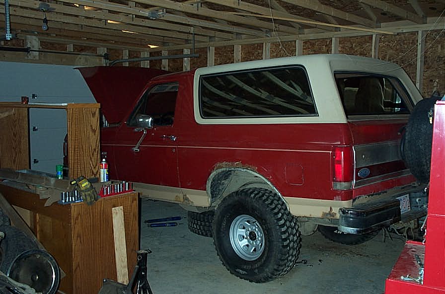Red and white 1987 Ford Bronco with lifted suspension and large tires, parked in a garage