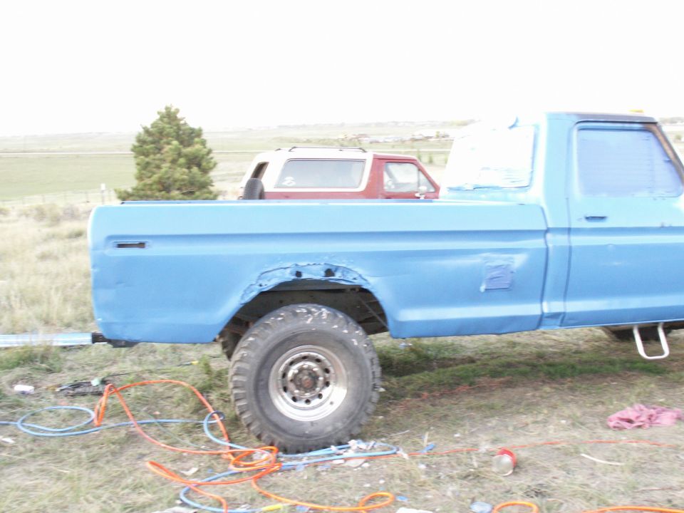 Blue pickup truck side view, showing rust damage on rear wheel well. A red SUV is in the bed