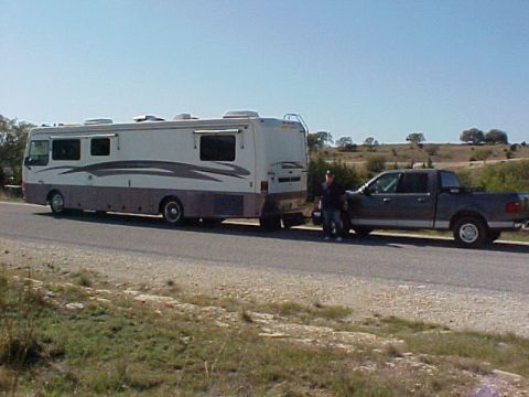 2002 Ford Supercrew - A gray pickup truck tows a large RV down a highway, with a person standing beside the truck.