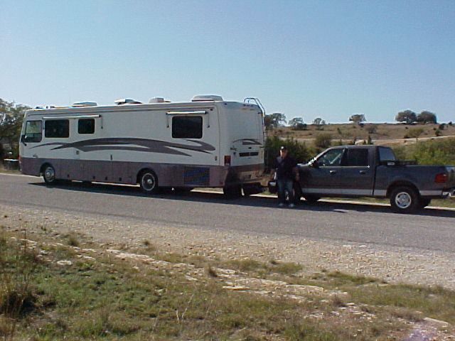 Dark Ford crew-cab pickup towed behind a tan Class A motorhome on a rural roadside