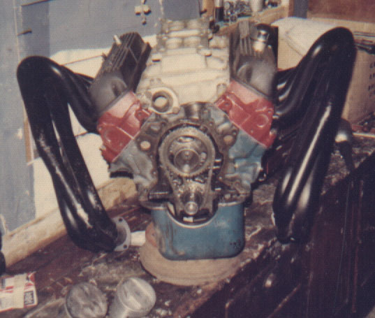 Engine block with red valve covers, black exhaust headers, and exposed timing gears, resting on a wooden surface