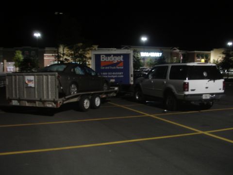 1989 Ford Bronco - A white SUV and a car on a trailer are parked in a lot at night, with a Budget truck nearby.
