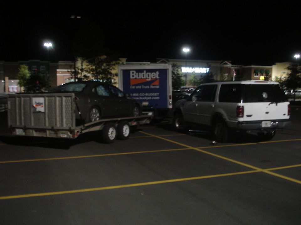 White SUV parked next to a car on a trailer, with a Budget truck behind them at night