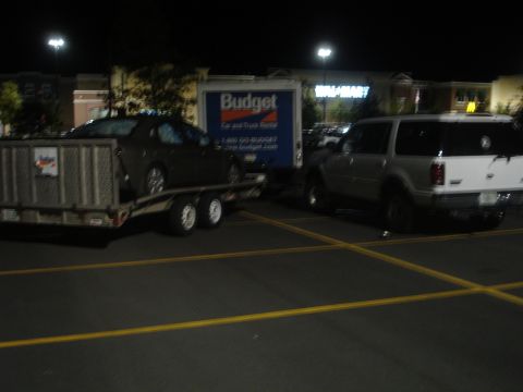 1989 Ford Bronco - A car is being towed on a trailer by a Budget rental truck at night in a parking lot at night.