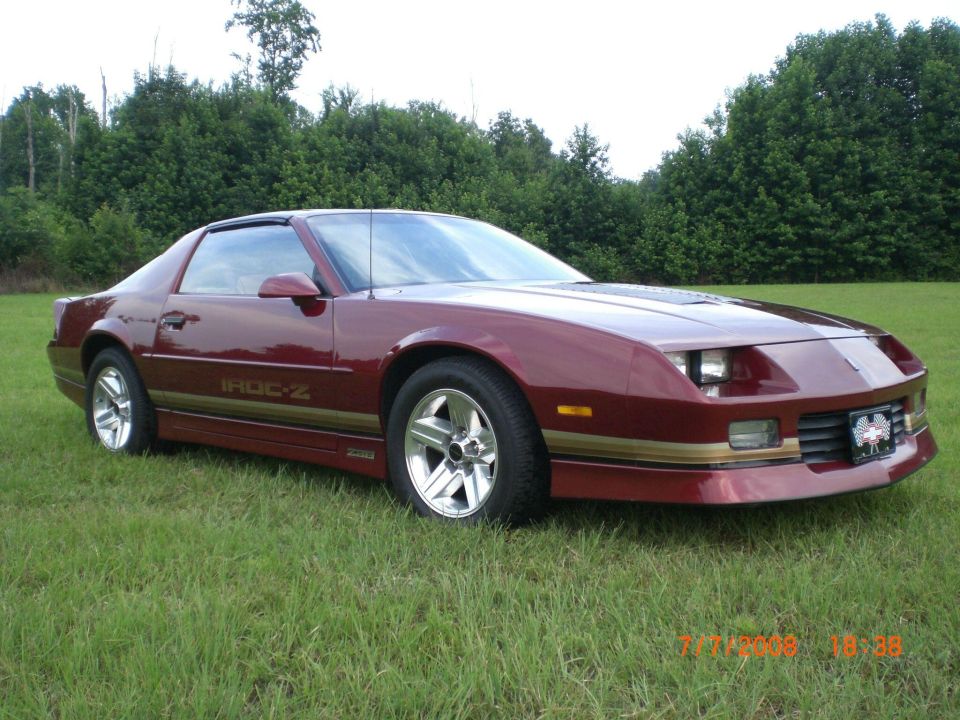 Maroon Chevrolet Camaro IROC-Z parked on grass, trees in background. "IROC-Z" decal visible on door
