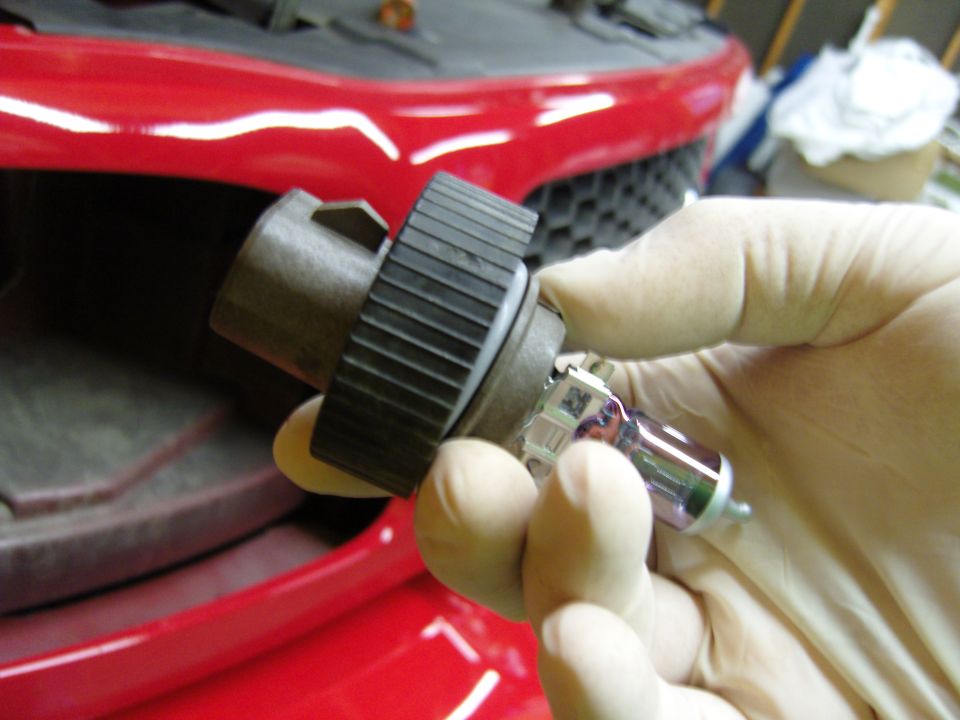 Gloved hands hold a headlight bulb with a black connector against a red car grille