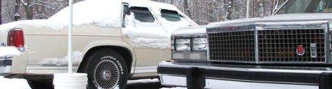 1991 Ford Crown Victoria LTD - A snow-covered beige sedan is parked next to another vehicle, with a white bucket visible in the foreground.