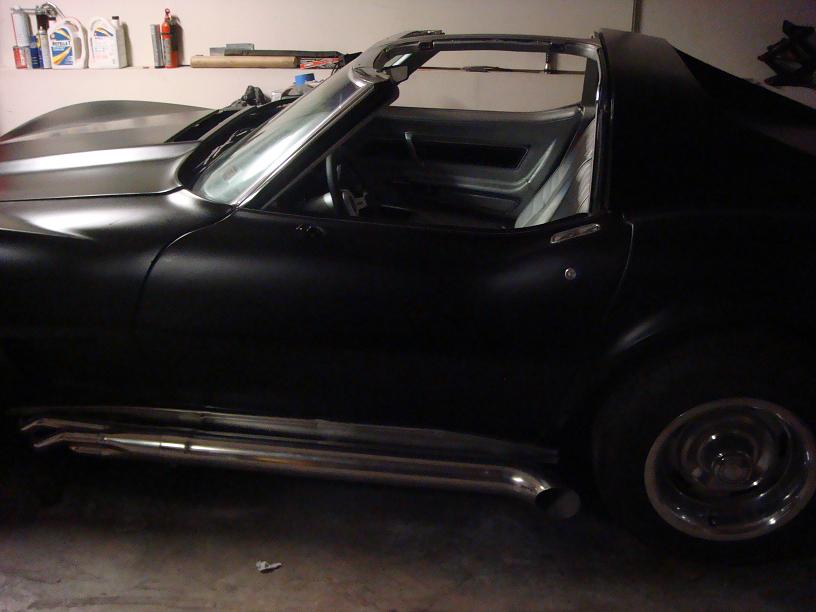 Side view of a black sports car with a matte finish and large chrome side pipes. The t-top roof is removed