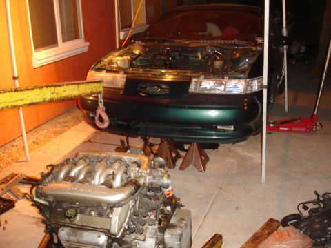 1993 Ford Taurus - The front of a green 1993 Ford Taurus is shown with its engine removed and placed on the ground in front of the vehicle.
