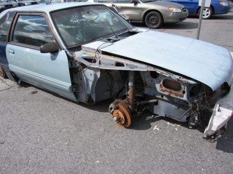 1991 Ford LTD Crown Victoria - A stripped-down Ford Mustang sits in a parking lot, showing significant rust and missing parts.