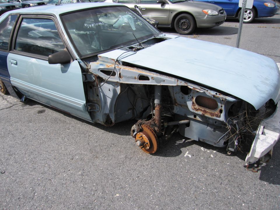 Light blue Ford Mustang coupe, missing front fender, headlight, and wheel. Blue door panel visible