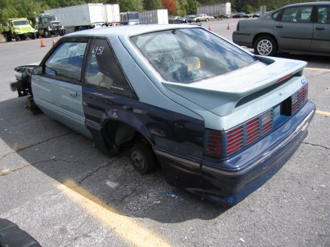 1991 Ford LTD Crown Victoria - A stripped-down Ford Mustang sits in a parking lot, missing its wheels and front bumper.