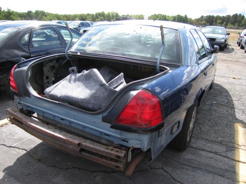 1991 Ford LTD Crown Victoria - A blue sedan sits in a junkyard with its trunk open, revealing disassembled parts and a missing bumper.