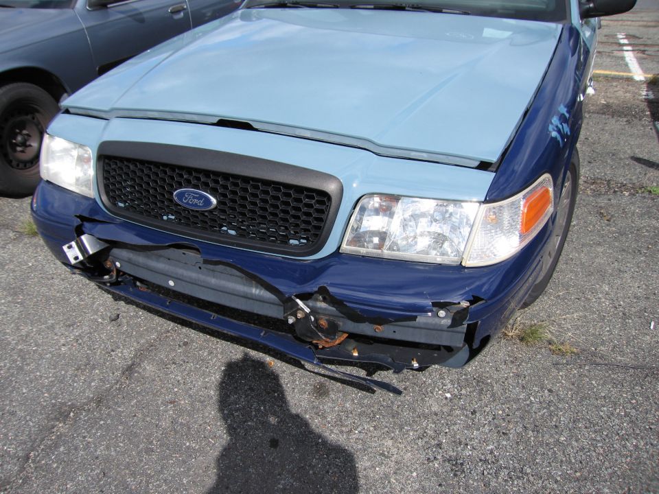 Front view of a damaged blue and light blue Ford Crown Victoria with a dented bumper and grille