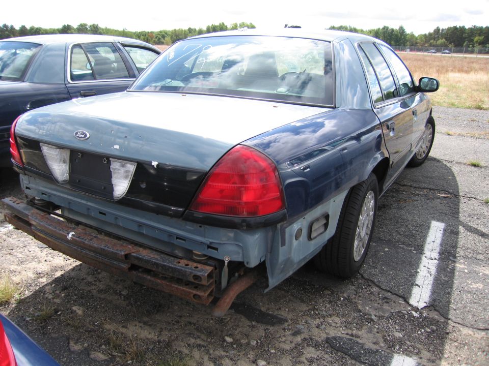 Rear view of a blue Ford Crown Victoria with missing bumper and body damage. Another car is parked behind it