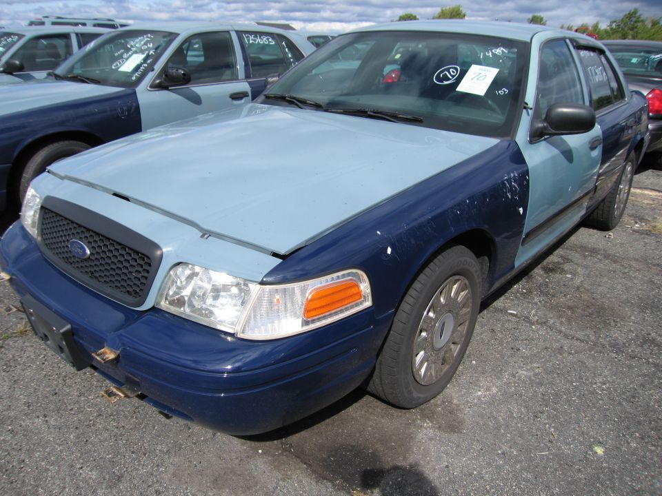 Blue and light blue Ford Crown Victoria with a black grille, visible damage on the front bumper, and a sticker on the windshield